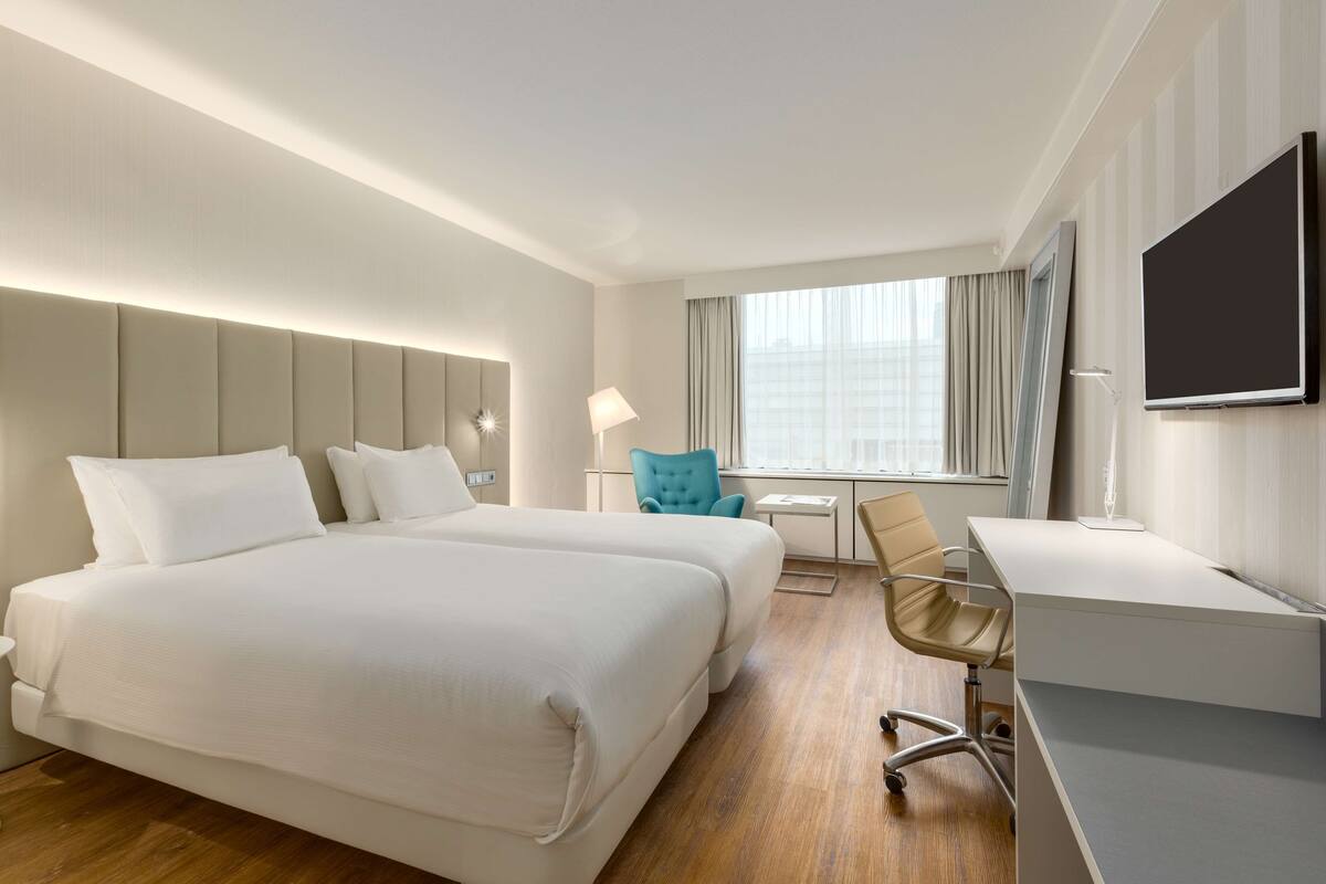 A hotel room with two beds, a desk, a chair, a TV, and a window with curtains.
