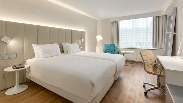 A hotel room with two beds, a desk, a chair, a small table, and a window with curtains.