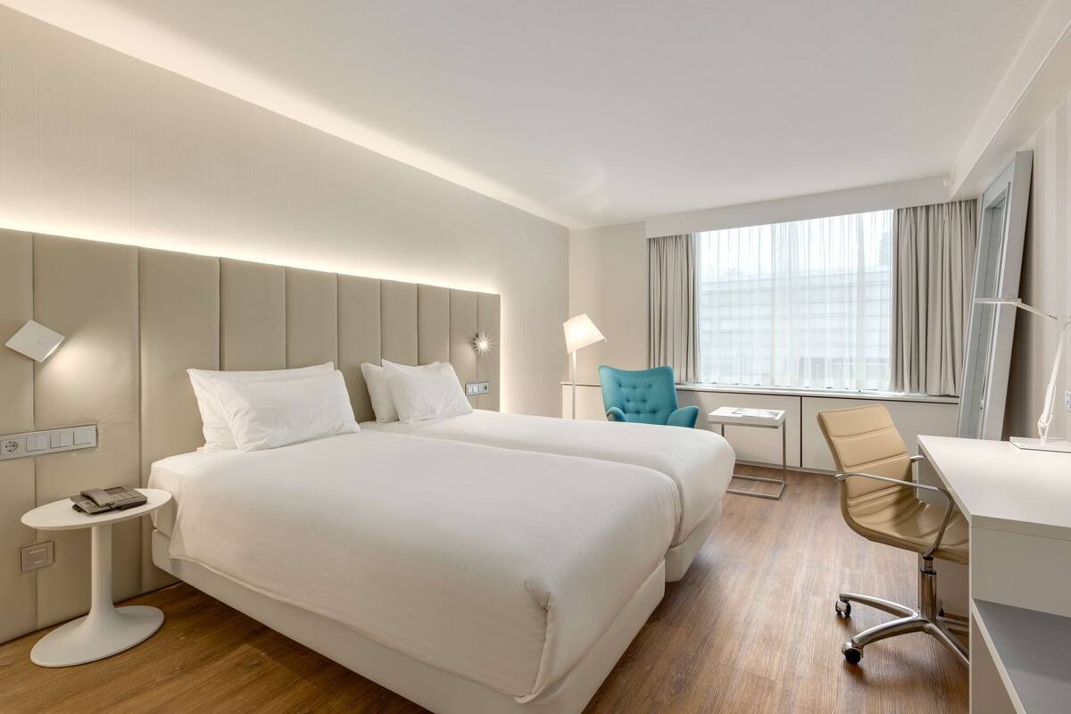 A hotel room with two beds, a desk, a chair, a small table, and a window with curtains.