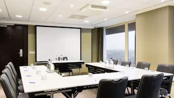 A conference room with a long table, chairs, a projector screen, and a city view through large windows.
