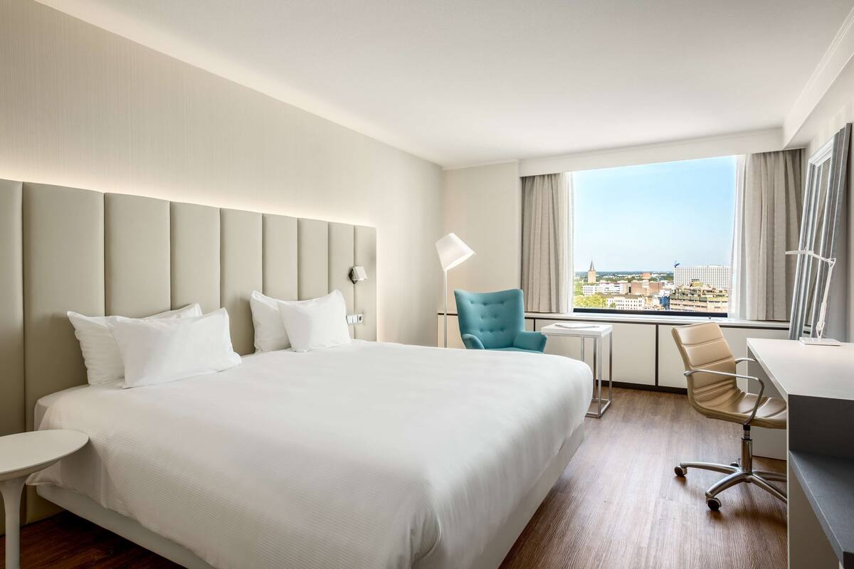 A hotel room with a large bed, a desk, a chair, and a view of the city.
