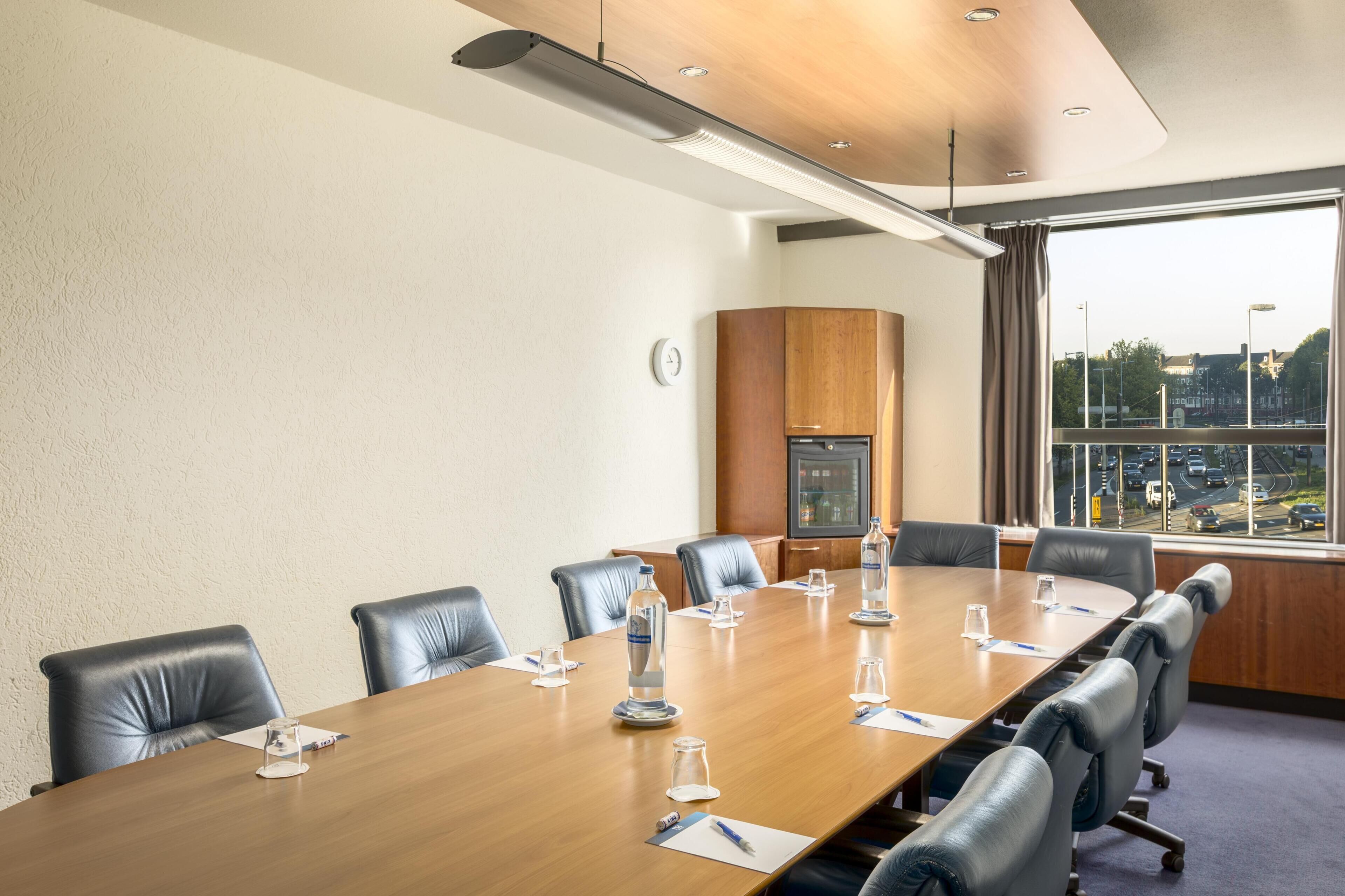 A conference room with a long wooden table, black leather chairs, and a view of a cityscape through large windows.