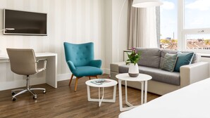 A modern hotel room with a flat-screen TV, a teal armchair, a grey sofa, and a small white table with a vase of flowers.