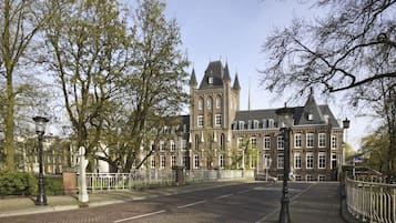 Un edificio histórico con una torre destacada y fachada simétrica, rodeado de un área pavimentada y árboles.