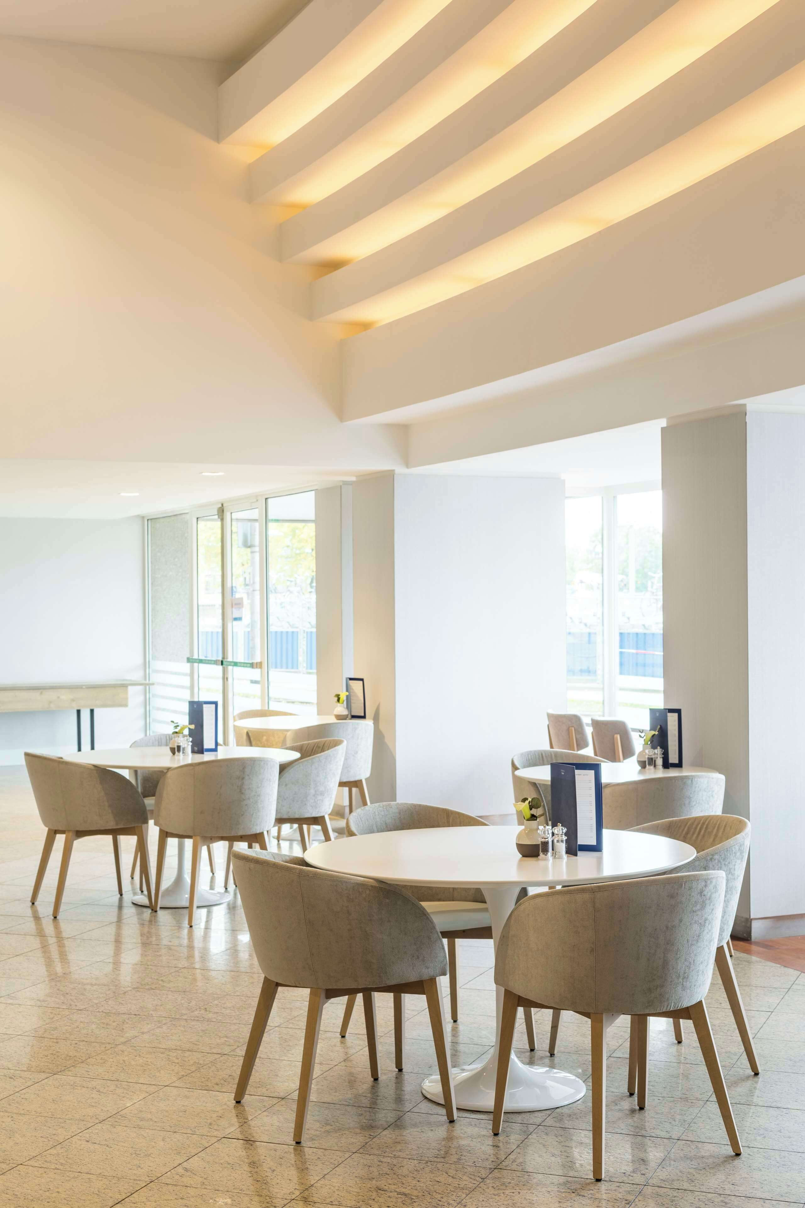 Un comedor moderno con mesas y sillas redondas, ventanas amplias y un techo alto con iluminación lineal.