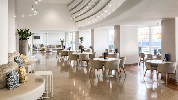 A spacious, modern lounge area with multiple seating arrangements, including sofas and round tables, and a curved ceiling with pendant lighting.