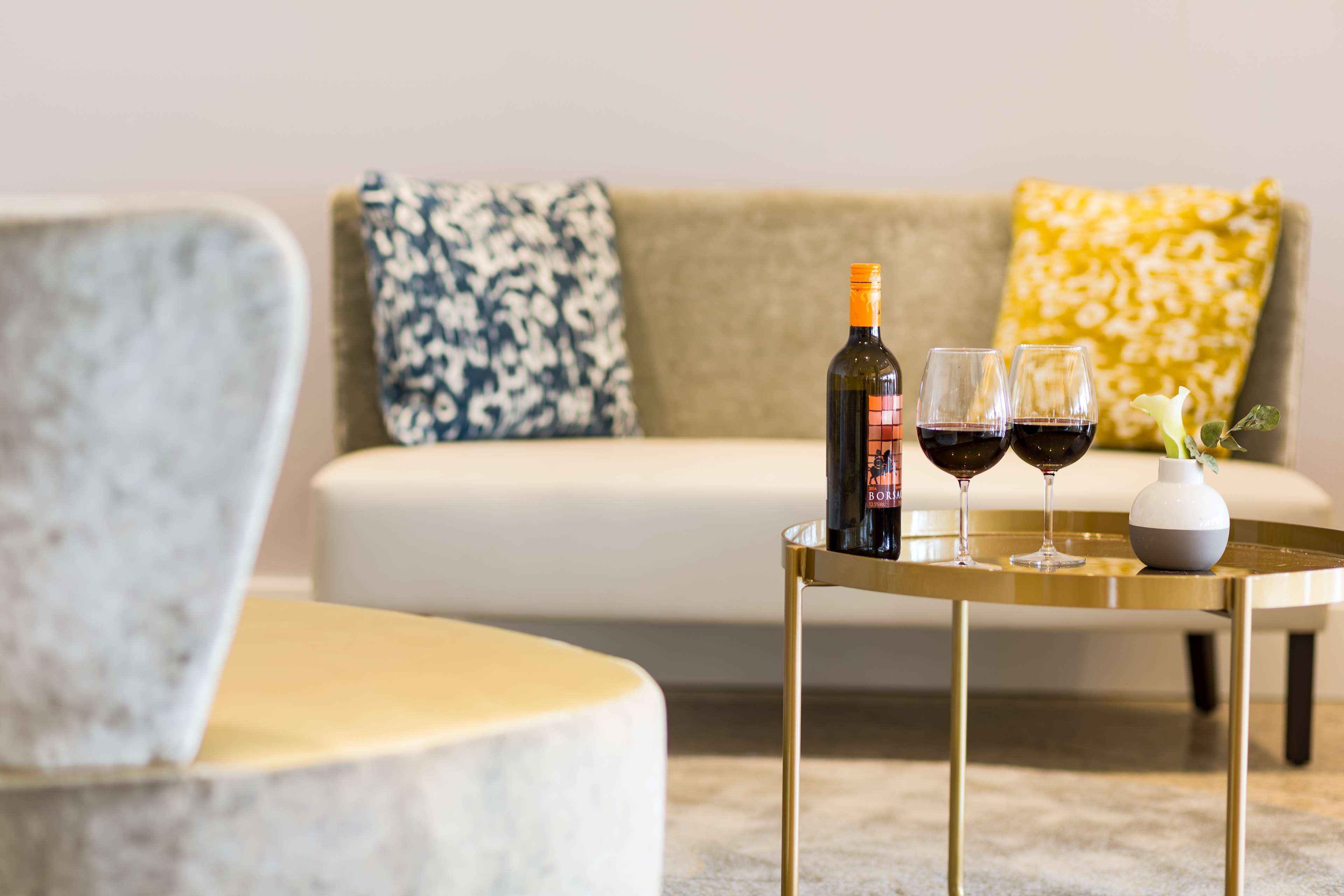 Un salon avec un canapé, une petite table ronde sur laquelle se trouvent une bouteille et des verres de vin, et un vase avec une fleur.