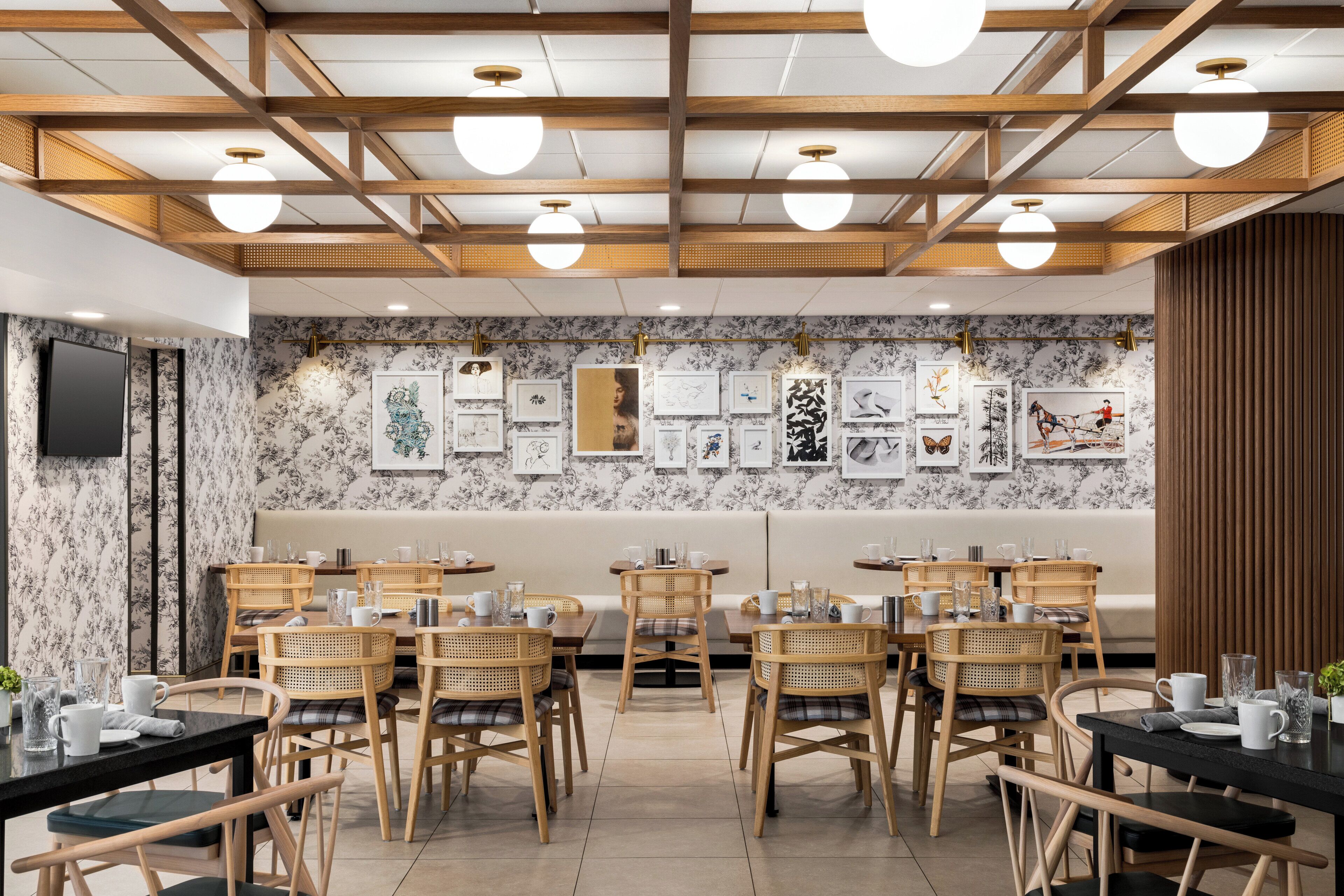 A restaurant with wooden ceiling beams, round ceiling lights, and a wall decorated with framed artwork. Tables are set with white plates, cups, and cutlery.
