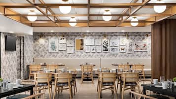 A restaurant with wooden ceiling beams, round ceiling lights, and a wall decorated with framed artwork. Tables are set with white plates, cups, and cutlery.