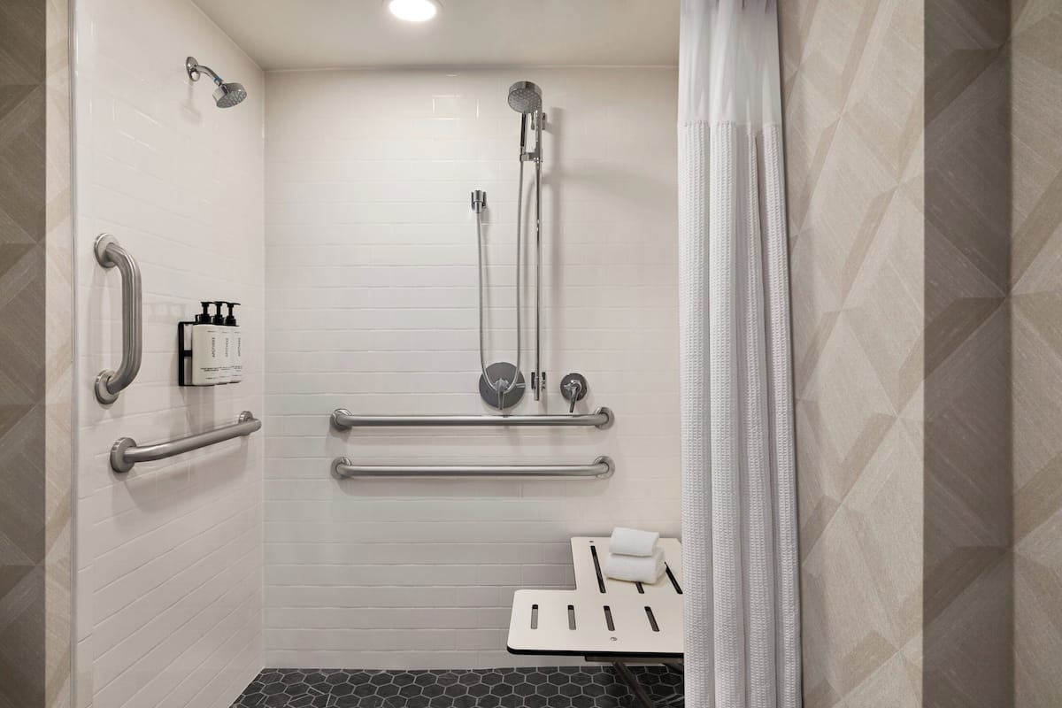 Une salle de bain moderne avec des murs carrelés de blanc, une douche équipée d’un pommeau de douche à main, de barres d’appui et de toilettes dotées d’un siège de sécurité.