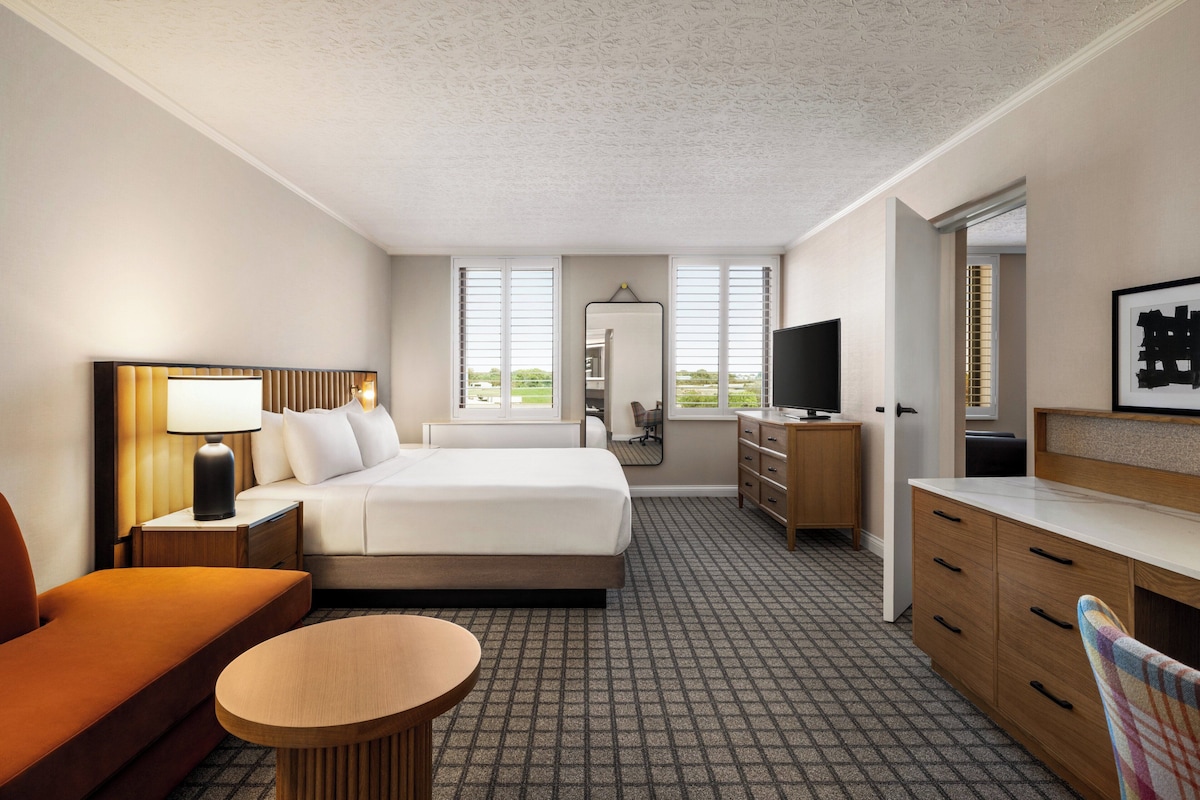 Ein Hotelzimmer mit Bett, Schreibtisch, Stuhl, Fernseher und einem Fenster mit Jalousien.