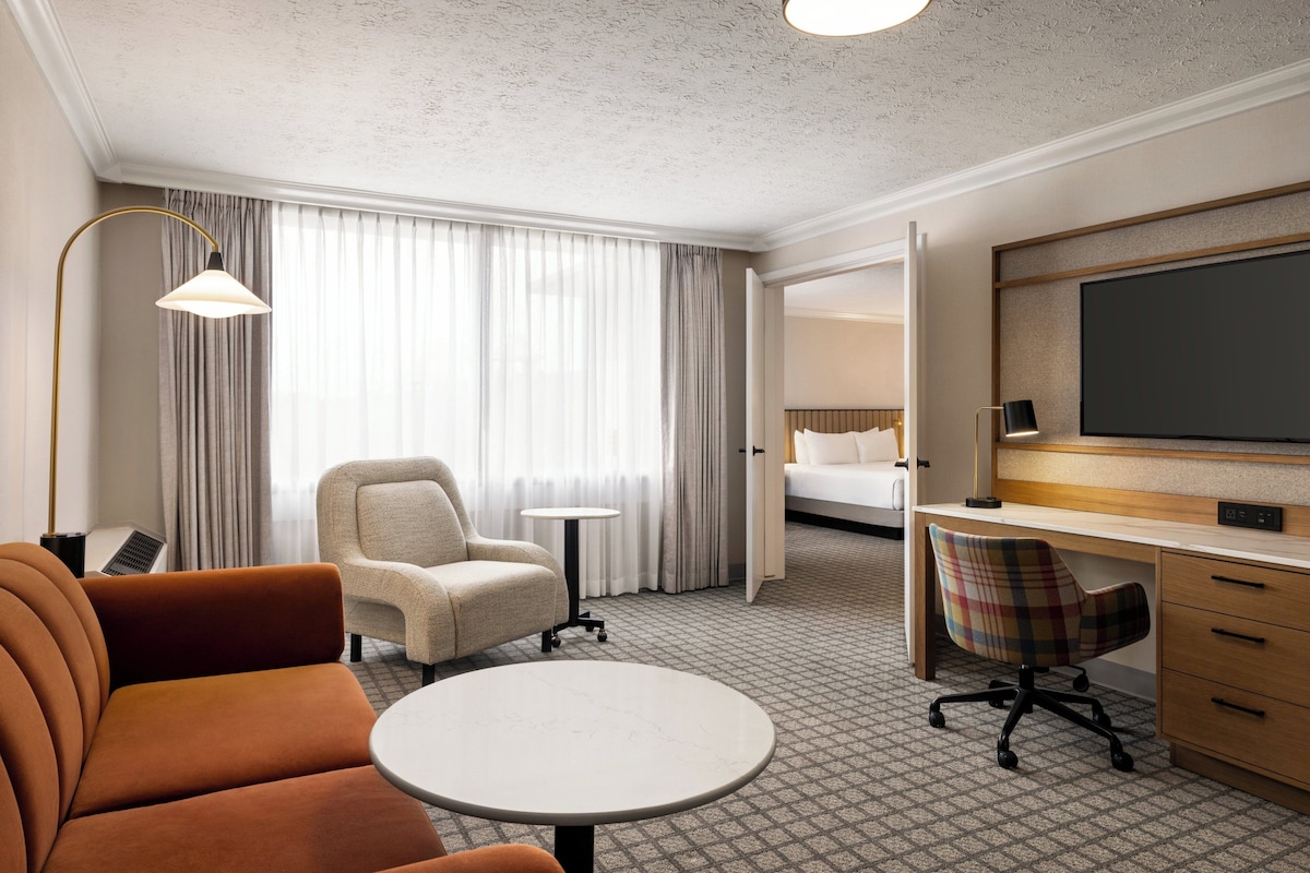 Ein Hotelzimmer mit einer Sitzecke, einem kleinen runden Tisch, einem Sessel, einem Schreibtisch, einem Fernseher und einem Bett im Hintergrund.