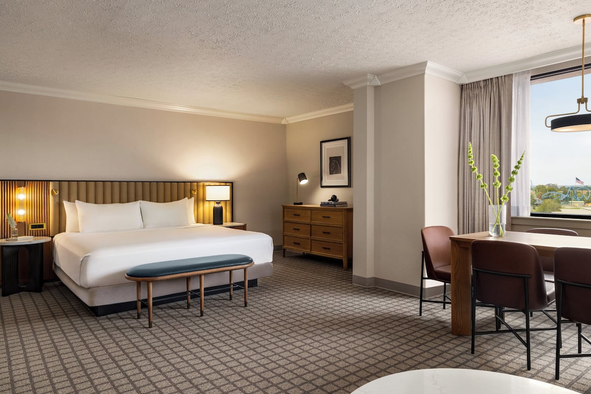 Une chambre d’hôtel avec un grand lit, un bureau avec des chaises, une commode et une fenêtre donnant sur un beau paysage.