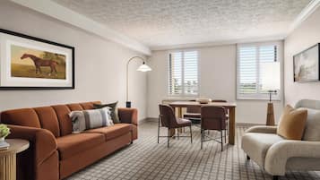 Un salón con un sofá marrón, un sillón beige, una mesa de comedor con sillas, una lámpara de pie y un cuadro de un caballo enmarcado en la pared.