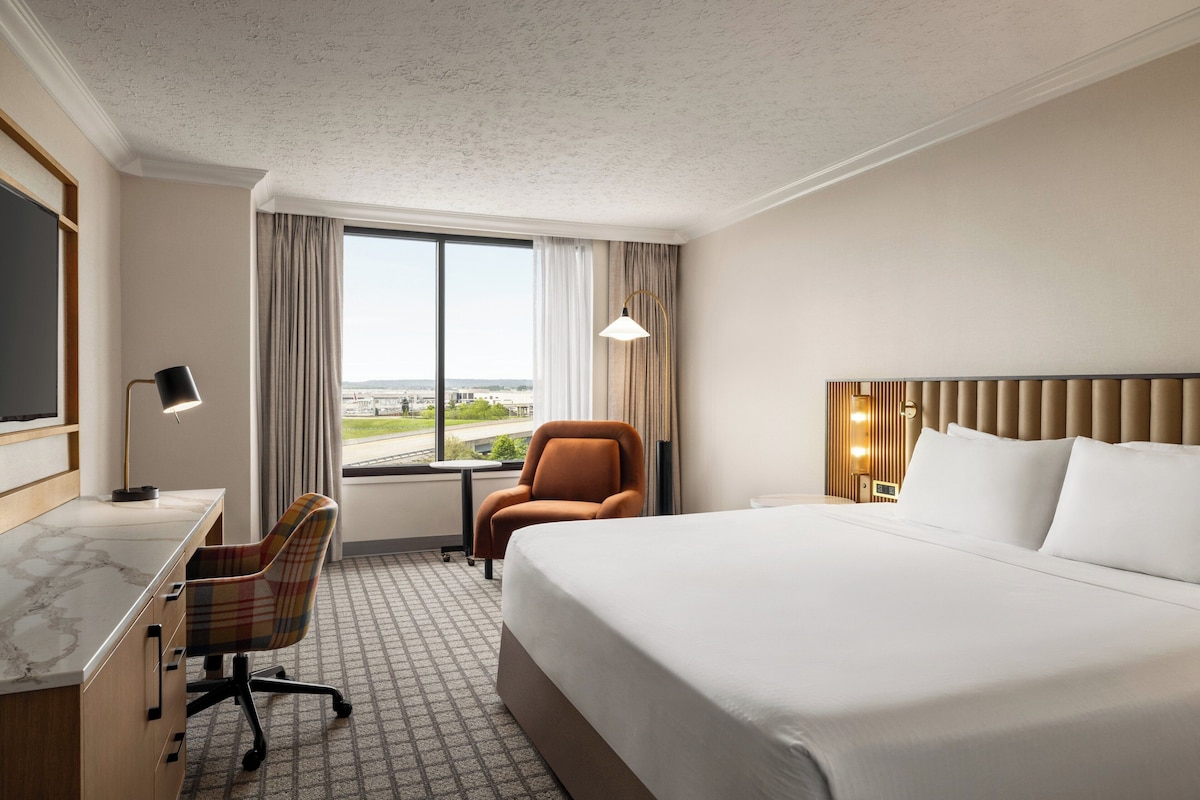 Ein Hotelzimmer mit einem großen Bett, einem Schreibtisch mit Stuhl, einem Fernseher und einem Fenster mit Vorhängen.