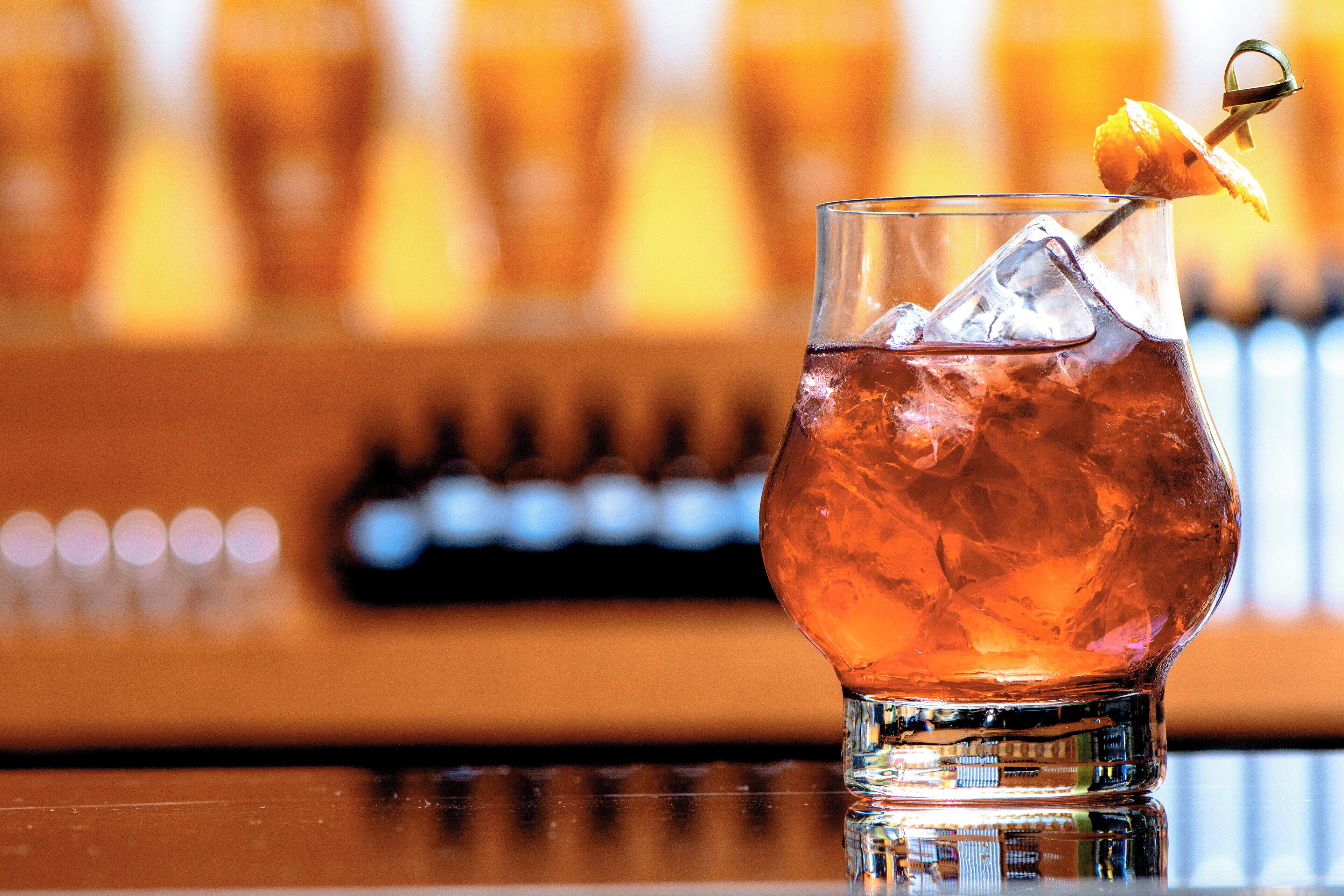 A glass of amber-colored beverage with ice cubes and a lemon garnish on a bar counter.