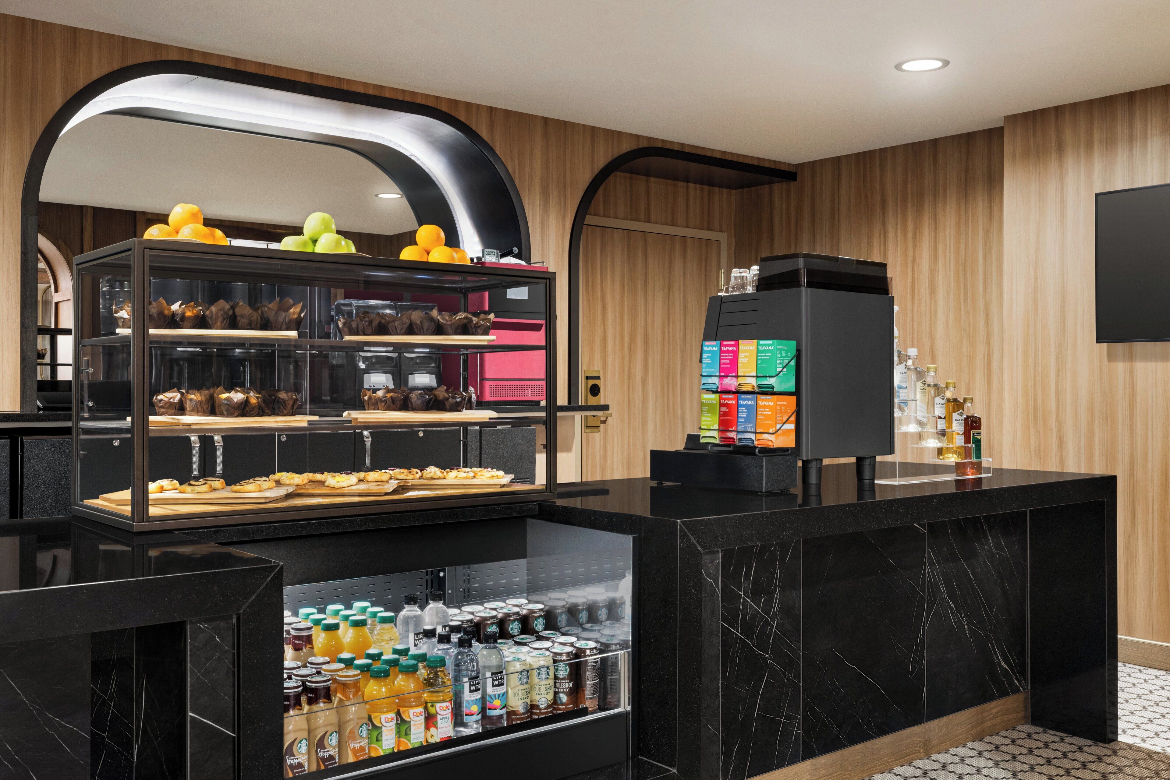 A modern hotel kitchen with a self-service breakfast area, including a refrigerator stocked with beverages and a display case with pastries and fruits.