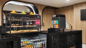 A modern hotel kitchen with a self-service breakfast area, including a refrigerator stocked with beverages and a display case with pastries and fruits.