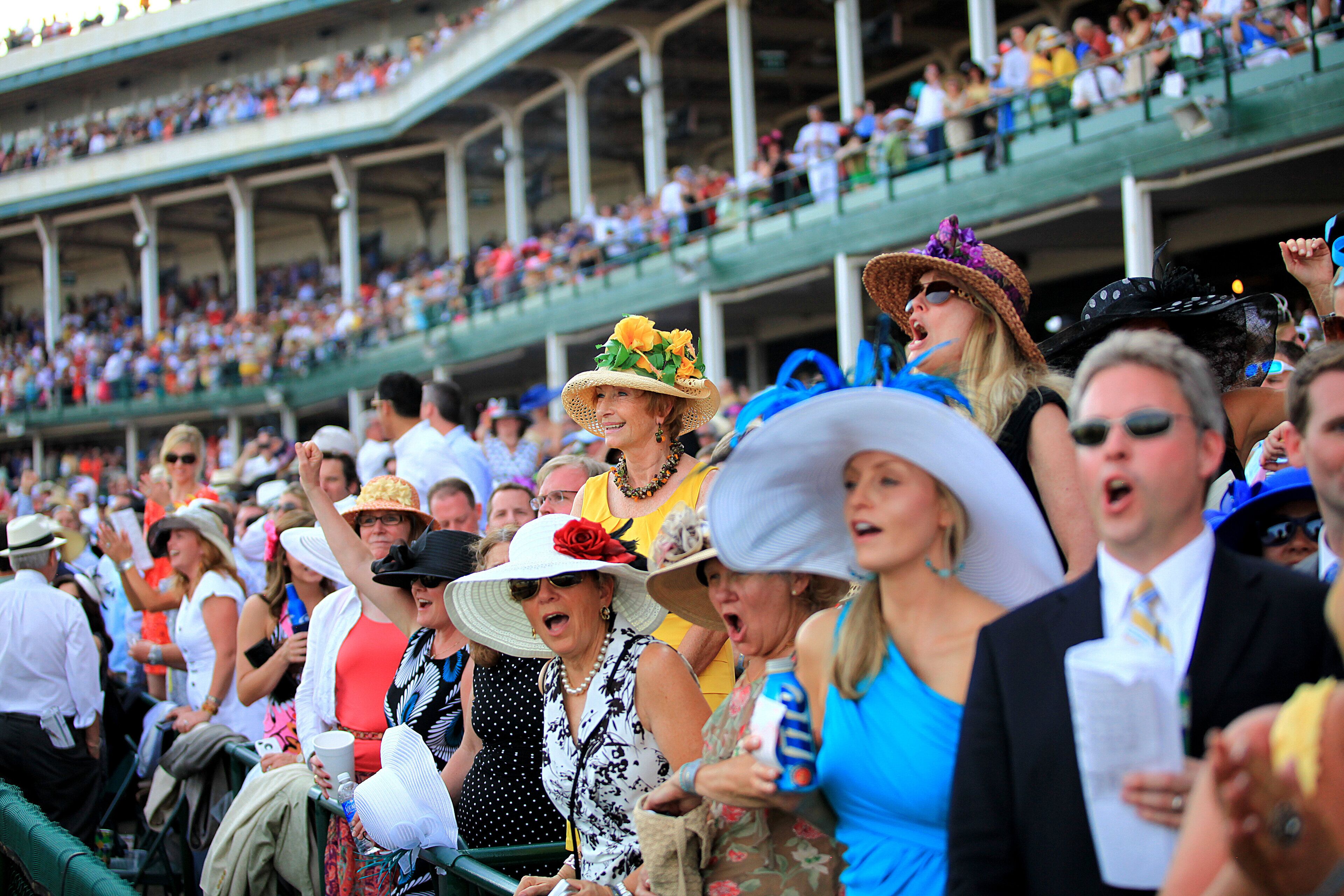 A crowded outdoor event with people standing and sitting, some wearing hats and others holding drinks.
