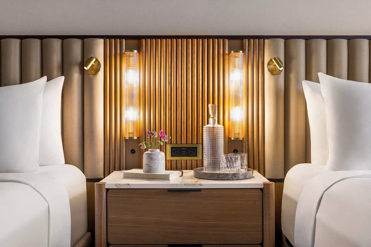 Une chambre d’hôtel avec deux lits, une tête de lit en bois, des lampes fixées au mur et une table de chevet agrémentée d’un vase et de verres.