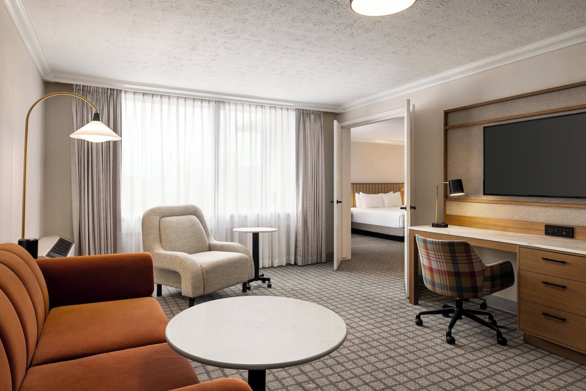 Une chambre d’hôtel avec un canapé, une petite table ronde, une chaise, un bureau, une télévision et un lit en arrière-plan.