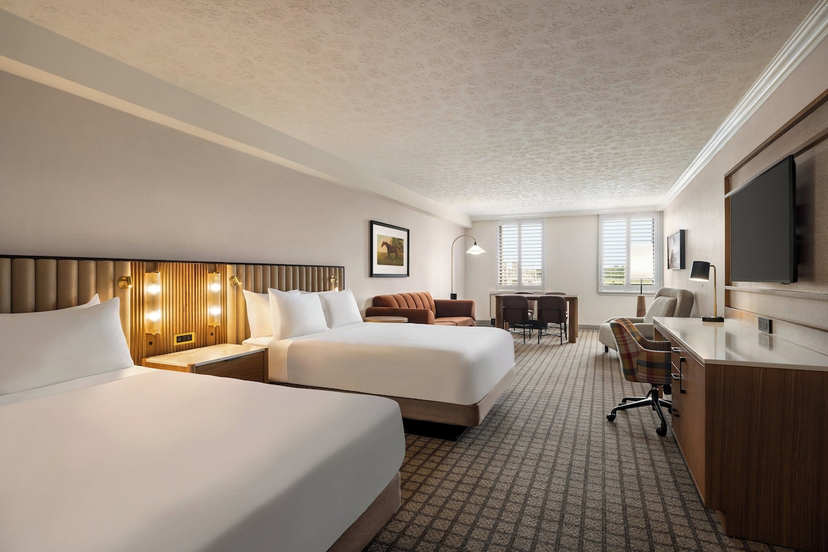 A hotel room with two beds, a desk, a chair, a TV, and a window with blinds.