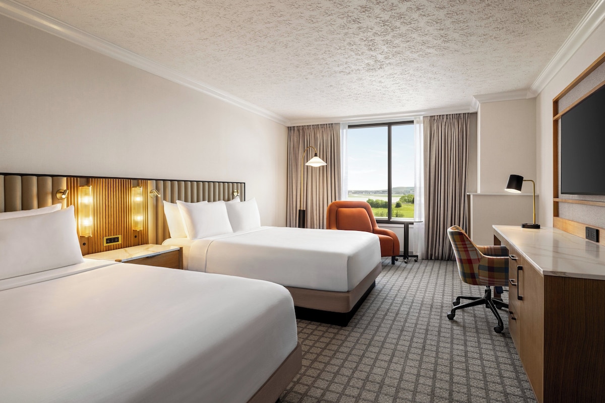 A hotel room with two beds, a desk, a chair, and a large window offering a view of the outdoors.