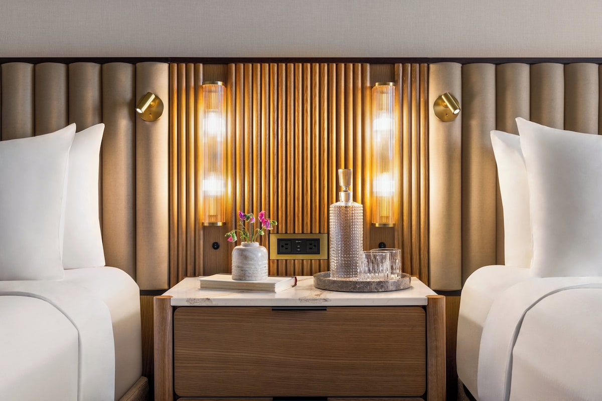 Ein Hotelzimmer mit zwei Betten, einem Holzkopfstück, Wandleuchten und einem Nachttisch mit Vase und Gläsern.