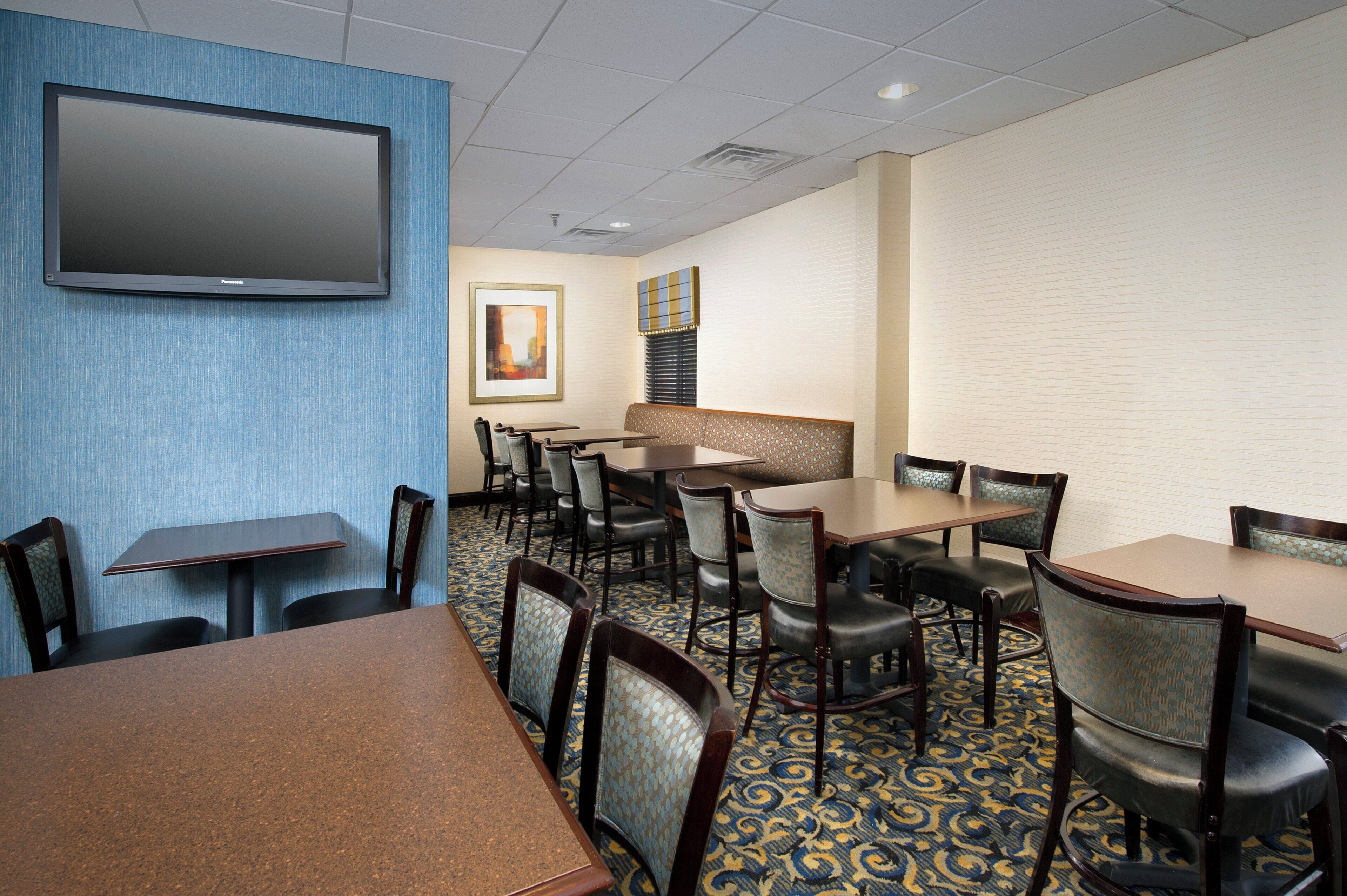 Een restaurantgedeelte in een hotel met meerdere tafels en stoelen, een wandgemonteerde flatscreen televisie en een kamerbreed tapijt met een patroon.