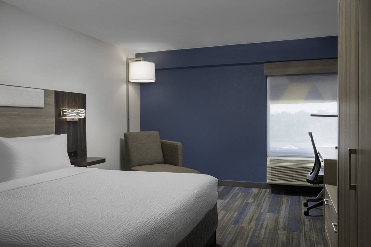 Une chambre d’hôtel avec un lit, une chaise, un bureau avec un ordinateur, une lampe et une fenêtre donnant sur un paysage agréable.