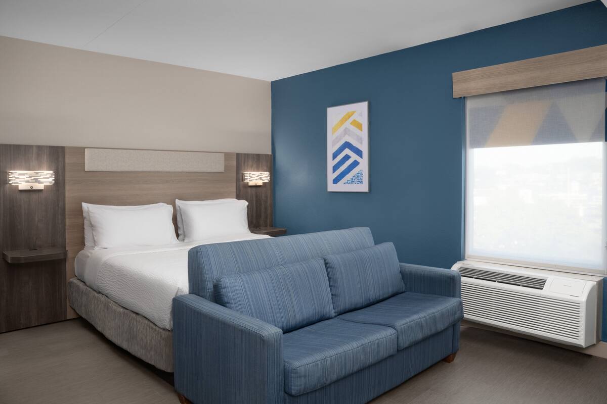 Une chambre d’hôtel moderne avec un lit, un canapé bleu, un climatiseur fixé au mur et une vue sur la ville par la fenêtre.