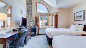 Habitación de hotel con pared de piedra, un amplio ventanal de arco, un escritorio con silla, dos camas y un televisor montado.