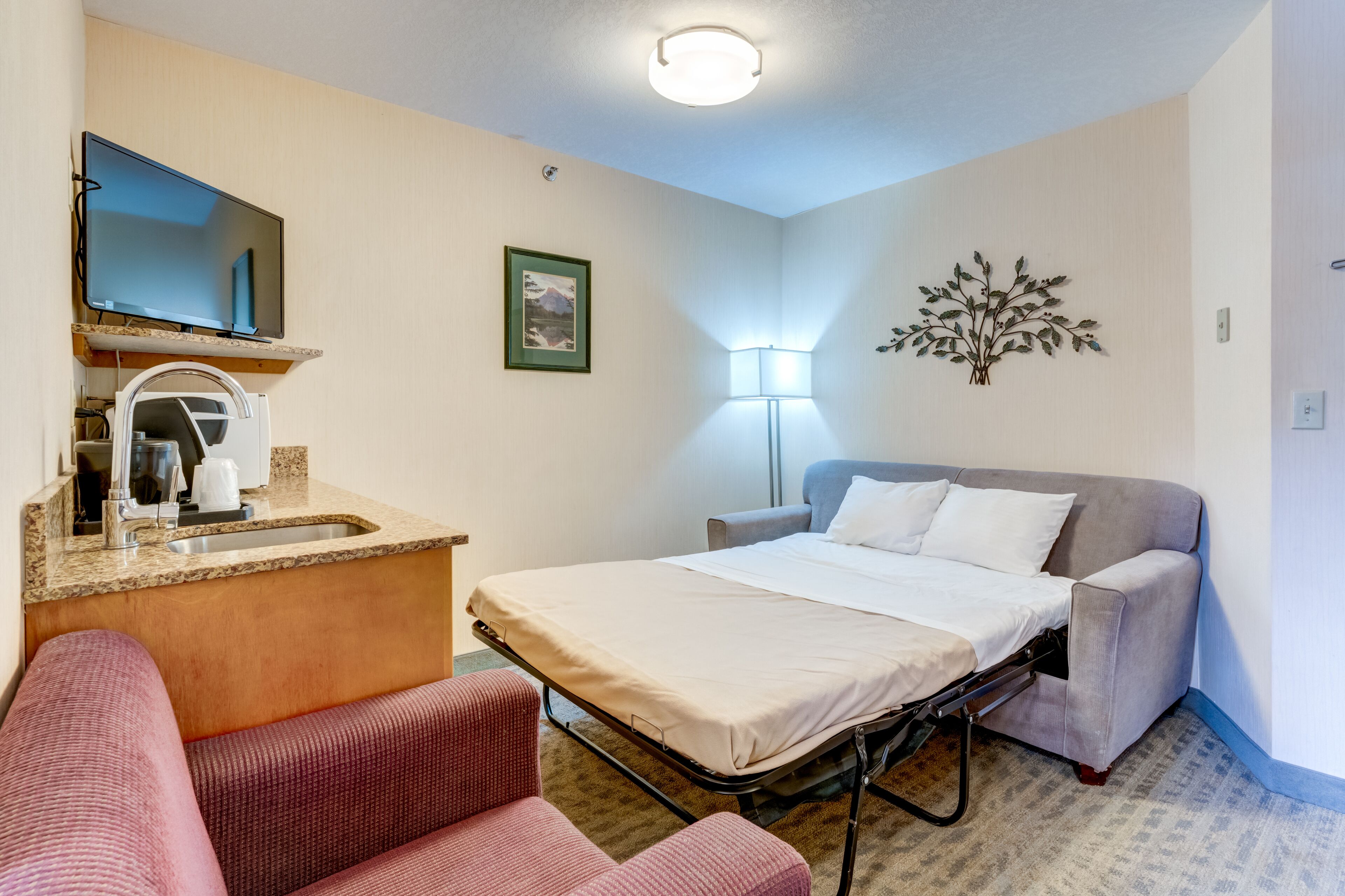 A hotel room with a bed, a desk with a sink, a chair, a lamp, and a wall-mounted decoration.