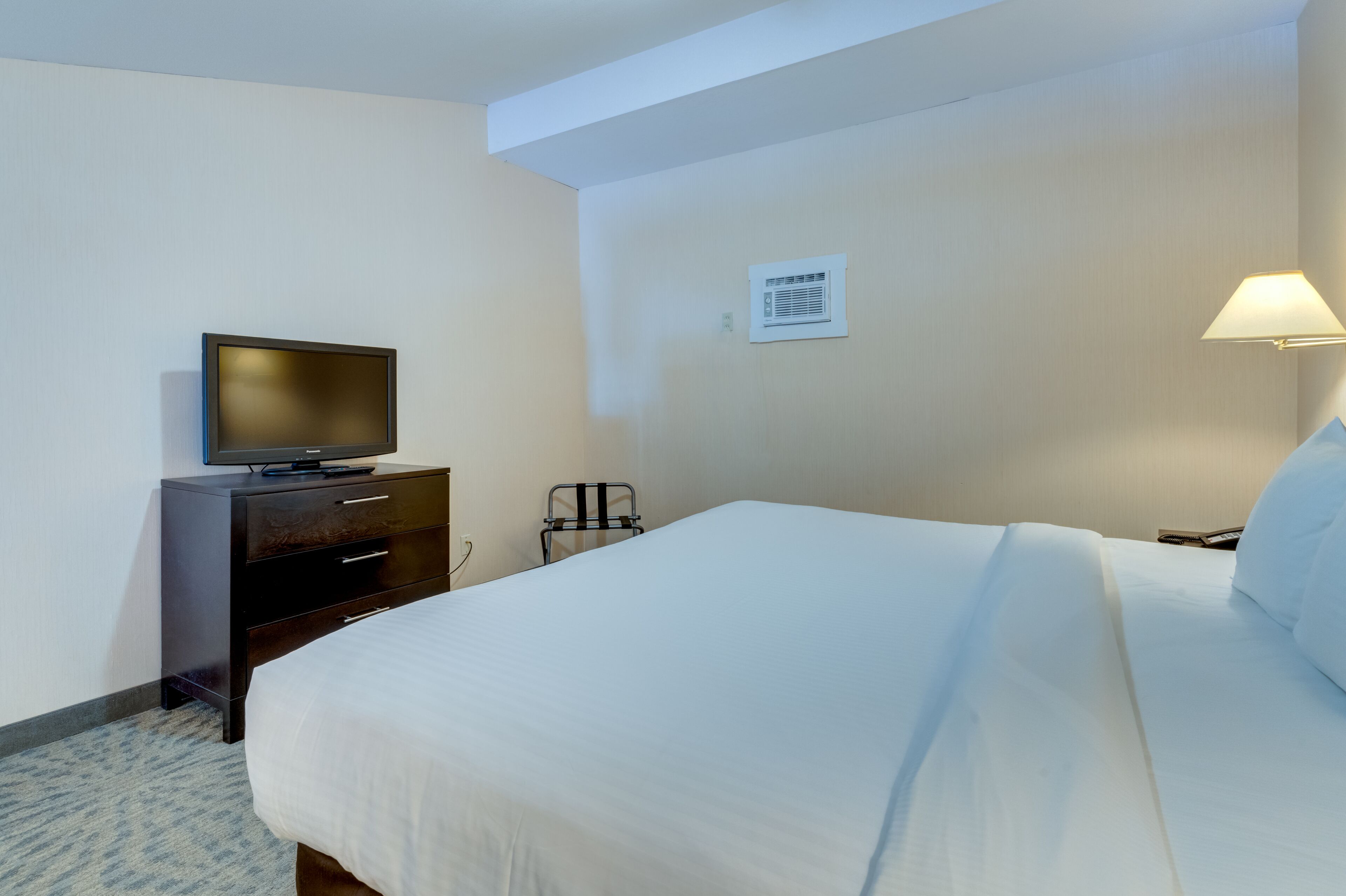 A hotel room with a bed, a television on a dresser, a lamp, and a wall-mounted air conditioning unit.