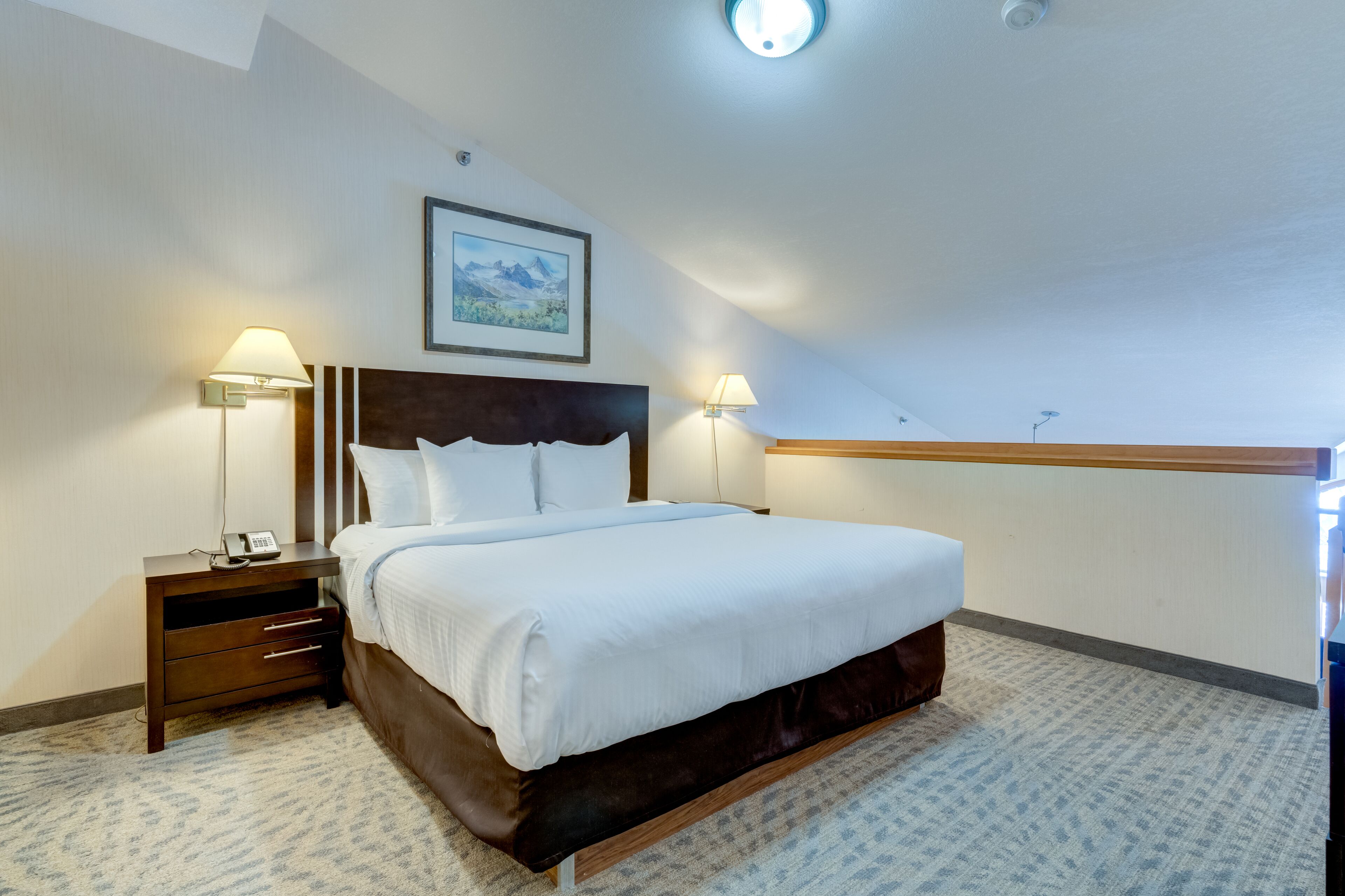 A hotel room with a large bed, two bedside tables with lamps, a framed picture on the wall, and a carpeted floor.