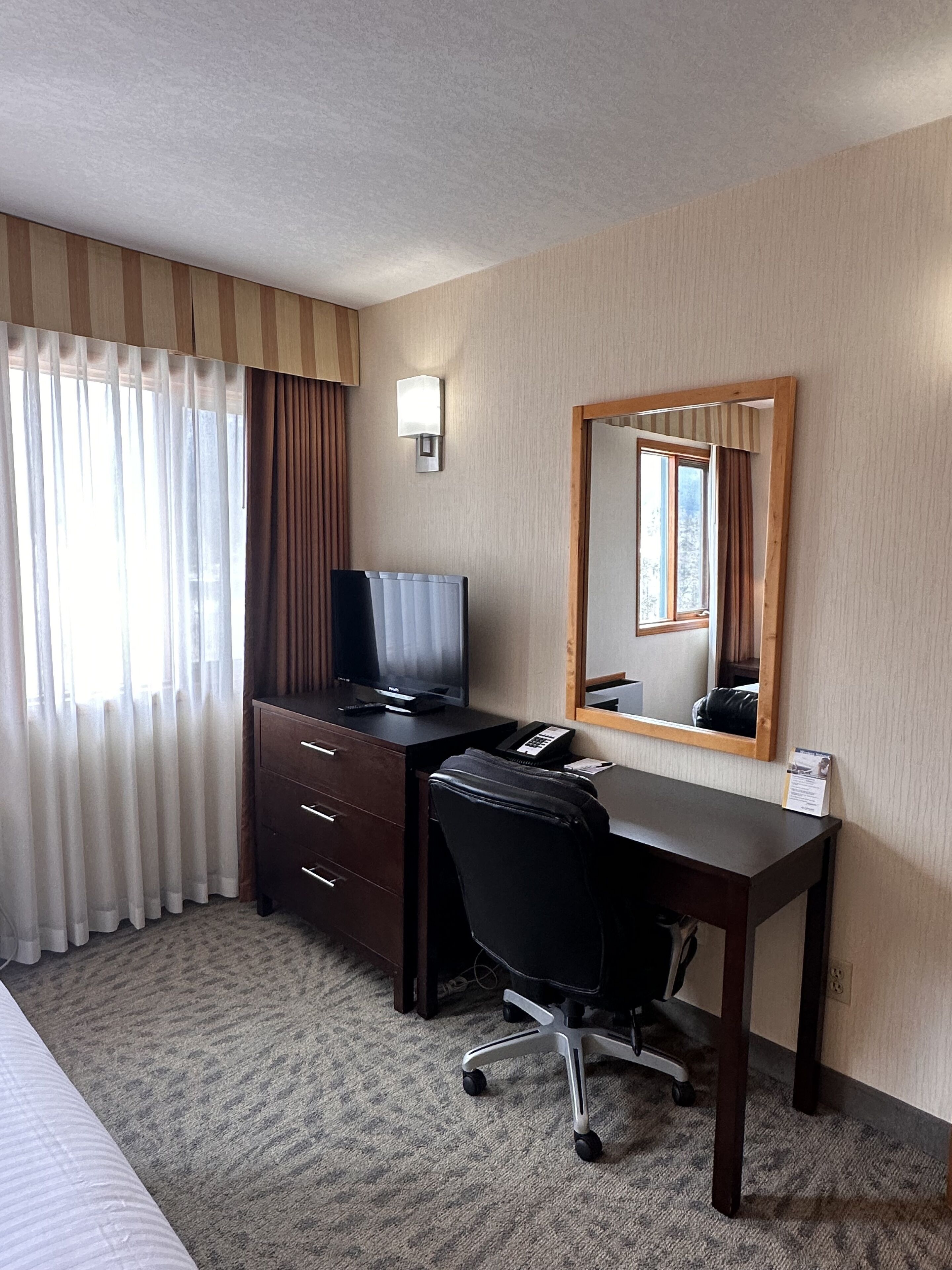 Une chambre d’hôtel équipée d’un bureau, d’une chaise, d’une télévision, d’un miroir et d’une fenêtre avec des rideaux.