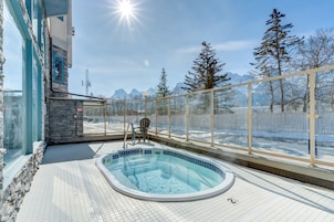 Área de piscina com estrutura de vidro, parede de pedra e vista para montanhas cobertas de neve.