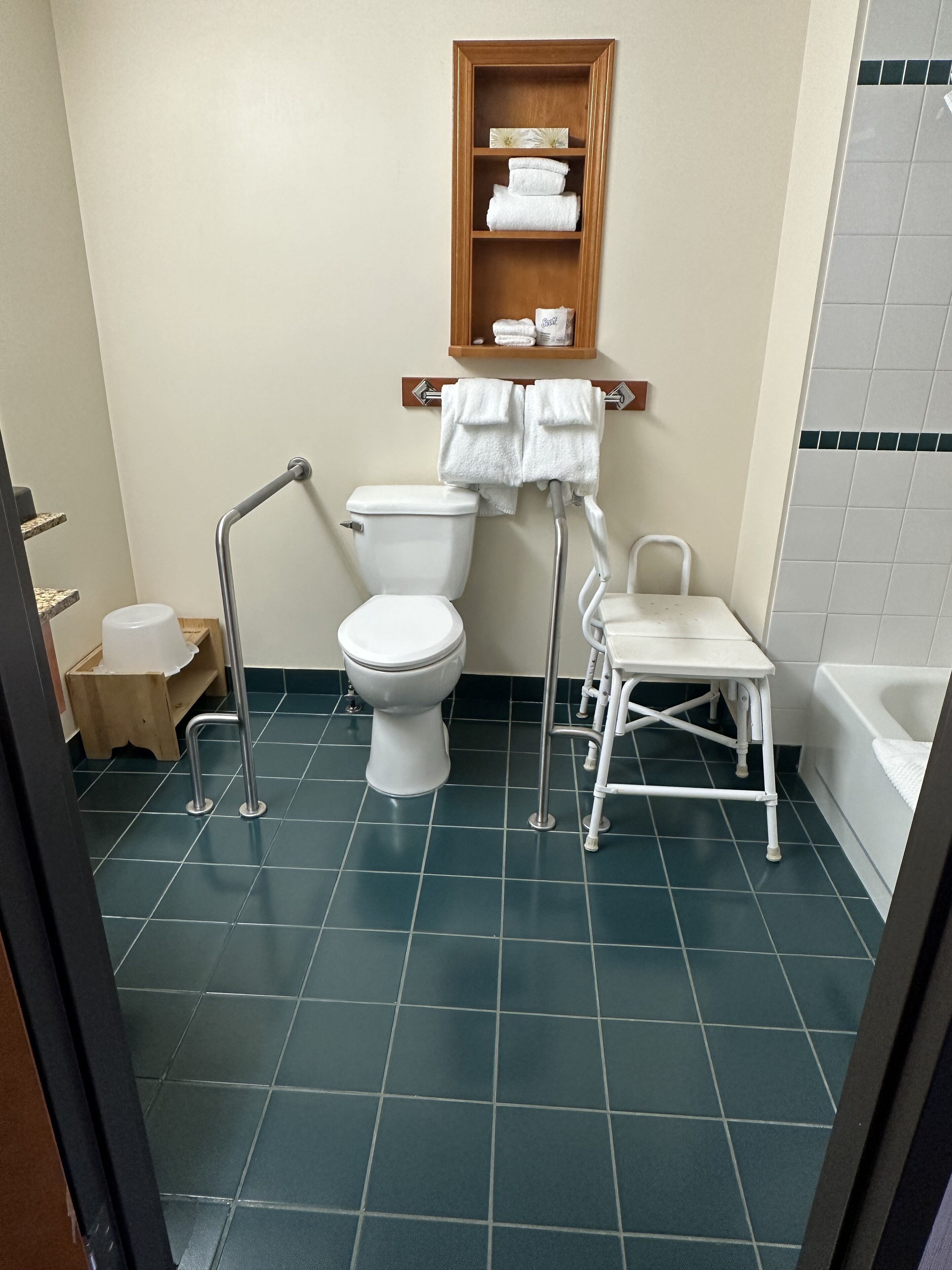 A bathroom with a white toilet, a wooden shelf above the toilet holding towels, a white chair, and a white bathtub.