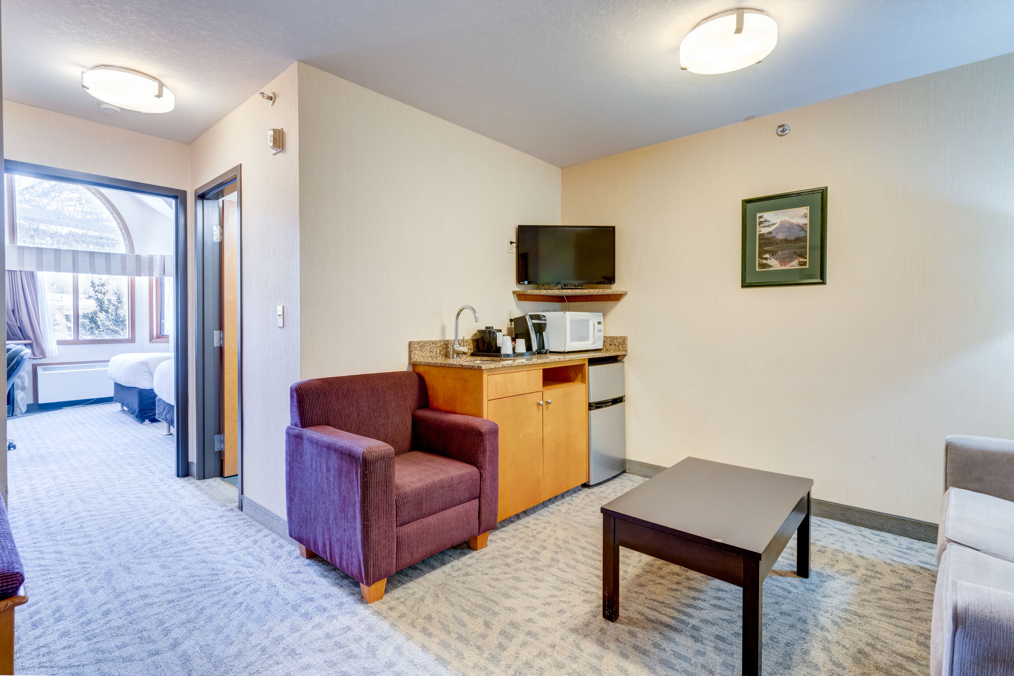 A hotel room with a kitchenette, a TV mounted on the wall, a framed picture, and a window with curtains.