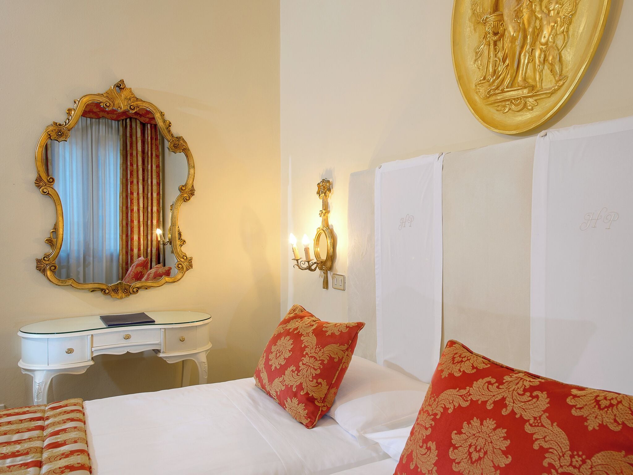 A hotel room with a large ornate mirror, a bedside table with a lamp, a bed with red and gold patterned pillows, and a wall-mounted artwork.