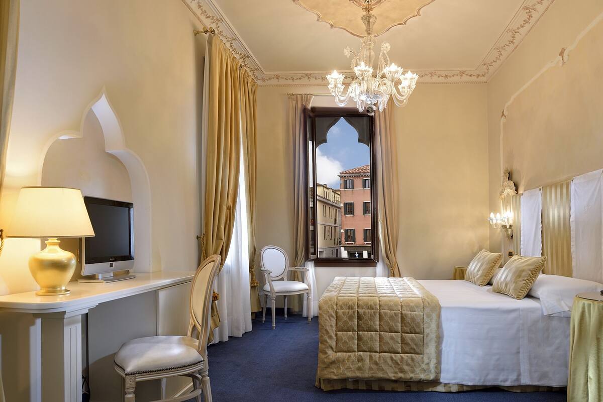 Une chambre d’hôtel avec un lit, un bureau doté d’une lampe, une chaise, un téléviseur et une vue sur les bâtiments par la fenêtre.