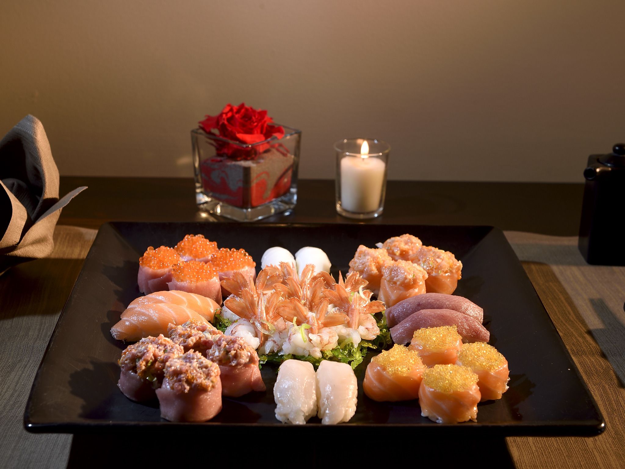 A black plate with various sushi pieces and a glass vase with a red rose in the background.