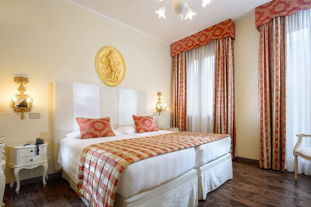 Une chambre d’hôtel avec un grand lit, deux tables de chevet, une petite table, un luminaire fixé au mur et une fenêtre avec des rideaux.