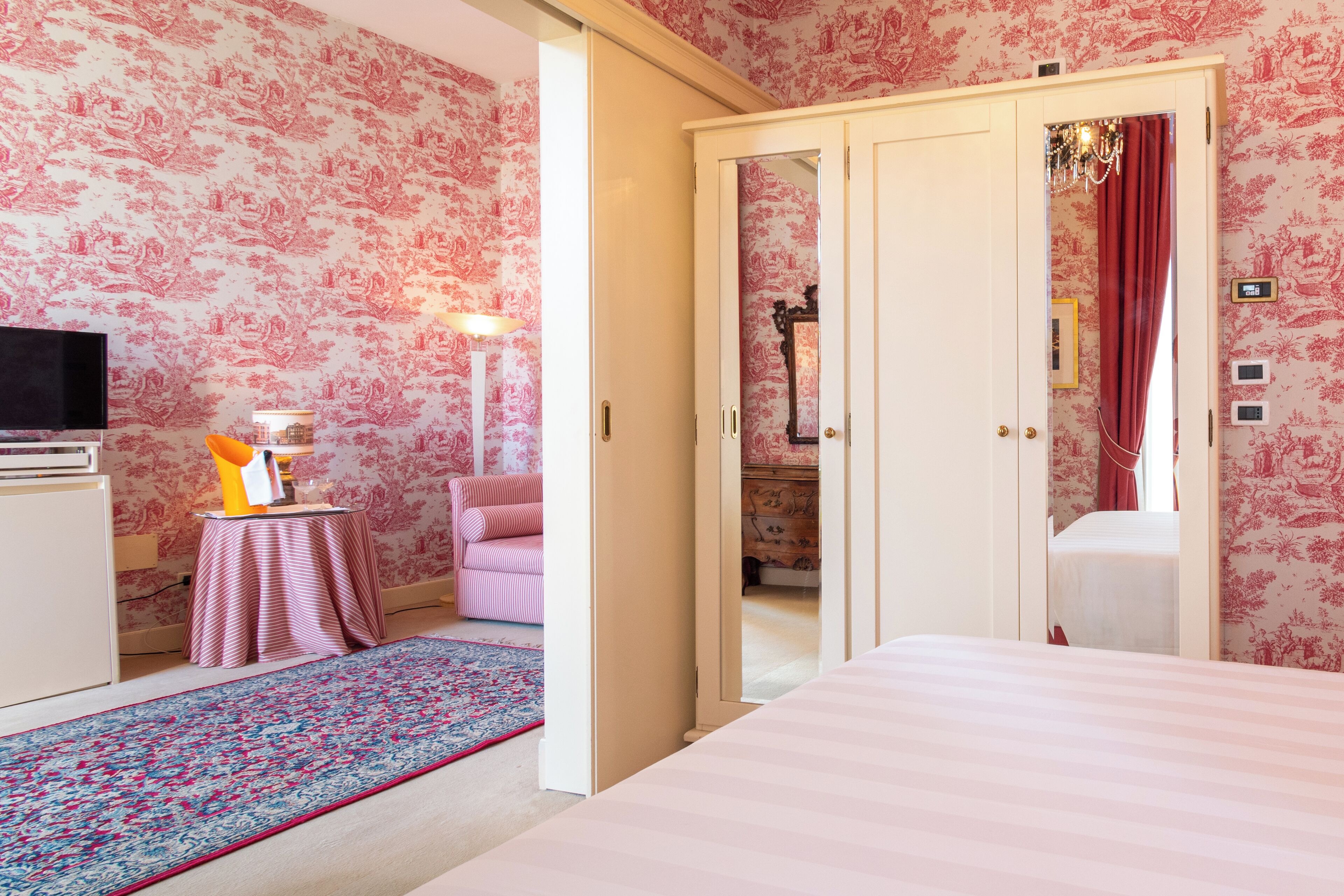 A room with floral wallpaper, a bed, a desk with a lamp, a chair, and a small table with a rug.