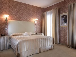Una camera d'albergo con un letto grande, due comodini, un tavolino, una lampada a parete, un quadro incorniciato e una finestra con tende.