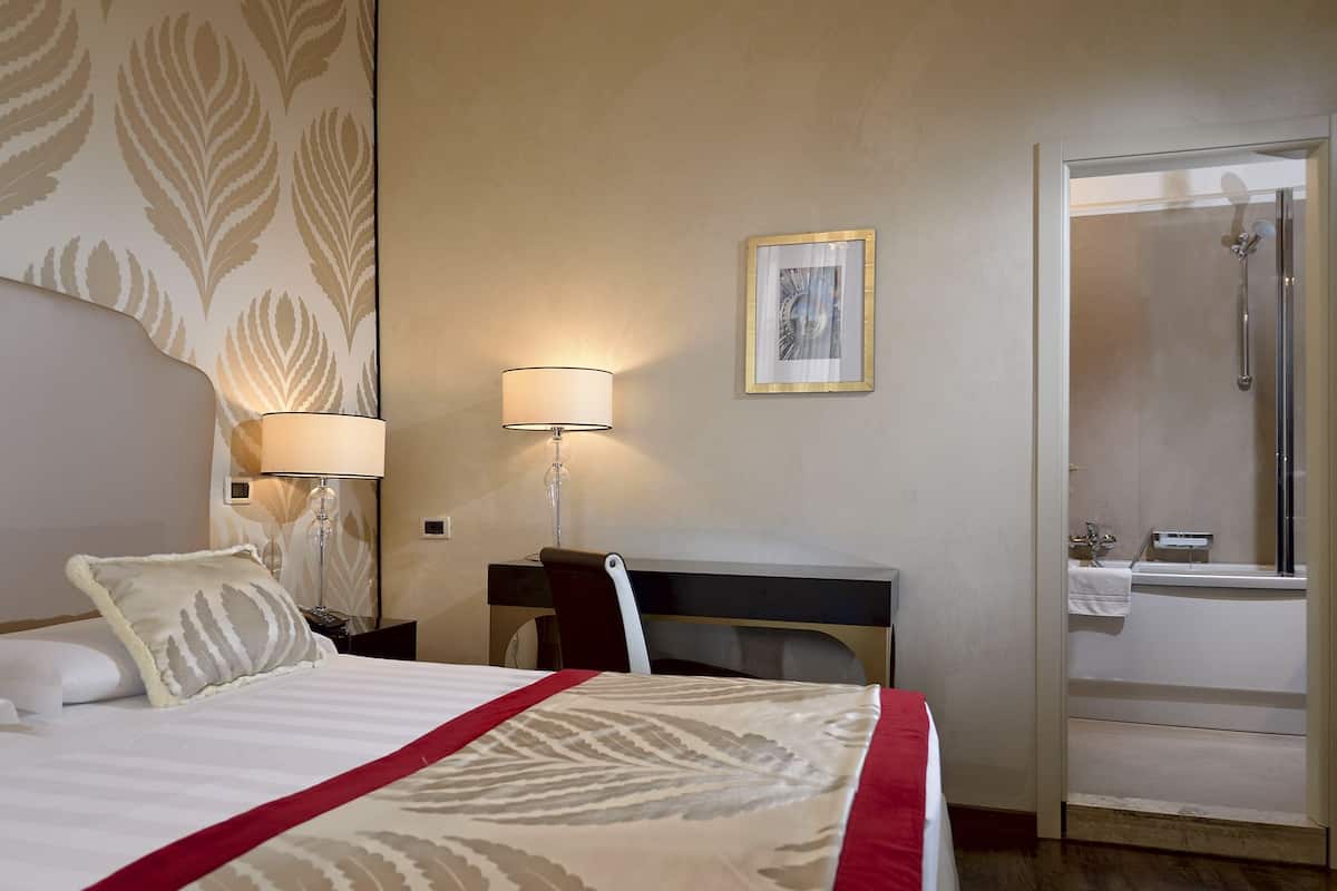 A hotel room with a bed, a desk, a chair, a lamp, a framed picture, and a bathroom with a shower.