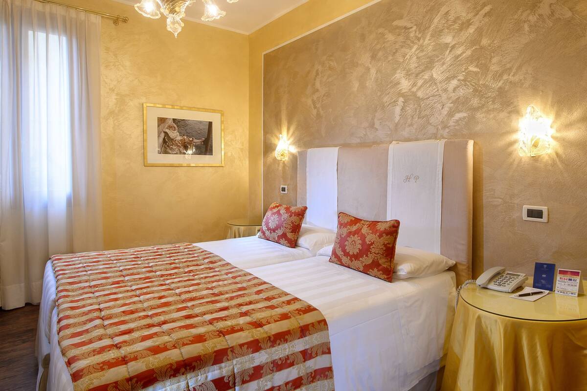 Une chambre d’hôtel avec un grand lit, deux oreillers, une table de chevet avec un téléphone et un panneau, et un lustre.