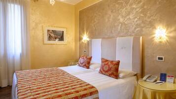 A hotel room with a large bed, two pillows, a bedside table with a phone and a sign, and a chandelier.
