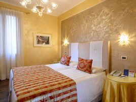 A hotel room with a large bed, two pillows, a bedside table with a phone and a sign, and a chandelier.