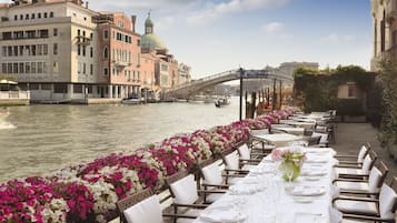 Un espace de restauration situé au bord de la rivière, avec des tables dressées pour le repas, offrant une vue sur un canal bordé de bâtiments historiques et d’un pont.
